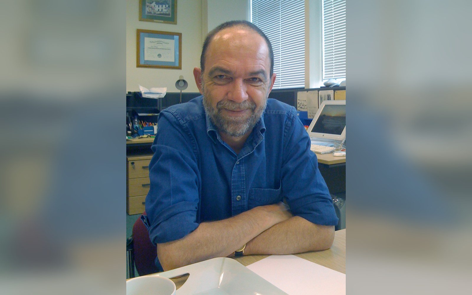 Bearded man in a blue shirt sits smiling with arms crossed at an office desk